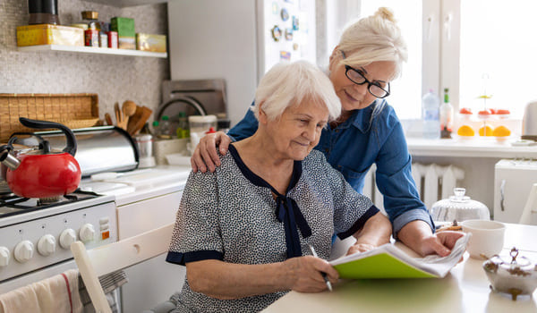 Eine Seniorin sitzt in der Küche und füllt Unterlagen aus, während ihre Tochter hinter ihr steht und sie dabei unterstützt.